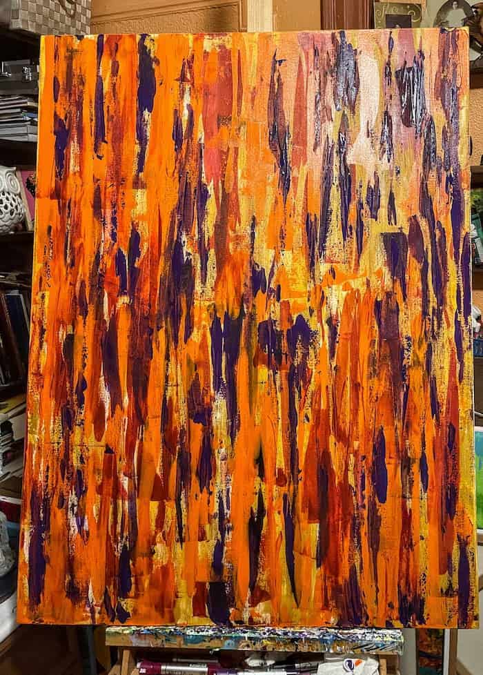an acrylic abstract painting on canvas made with orange, purple, yello,and red vertical paint strokes around the painting there are objects in tha background from the aetists studio such as an antique photo of a woman in a picture frame, paintbrushes, papers and other art materials